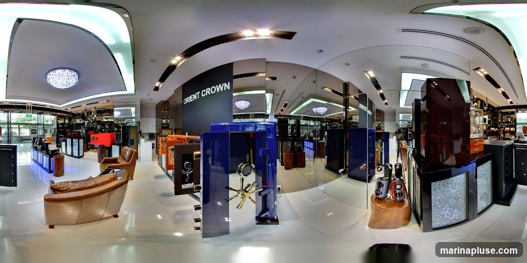 A panoramic view of a high-end showroom displaying various safes and luxury items. The store features a modern design with sleek displays, mirrored walls, and elegant lighting, including a large chandelier. Prominently displayed are several safes in different colors and sizes, including a striking blue safe with its door open, revealing its interior mechanism. Leather seating is arranged for customers. In the background, shelves are stocked with what appear to be watches or other valuable accessories. The store's name, "ORIENT CROWN," is visible on a dark wall.
