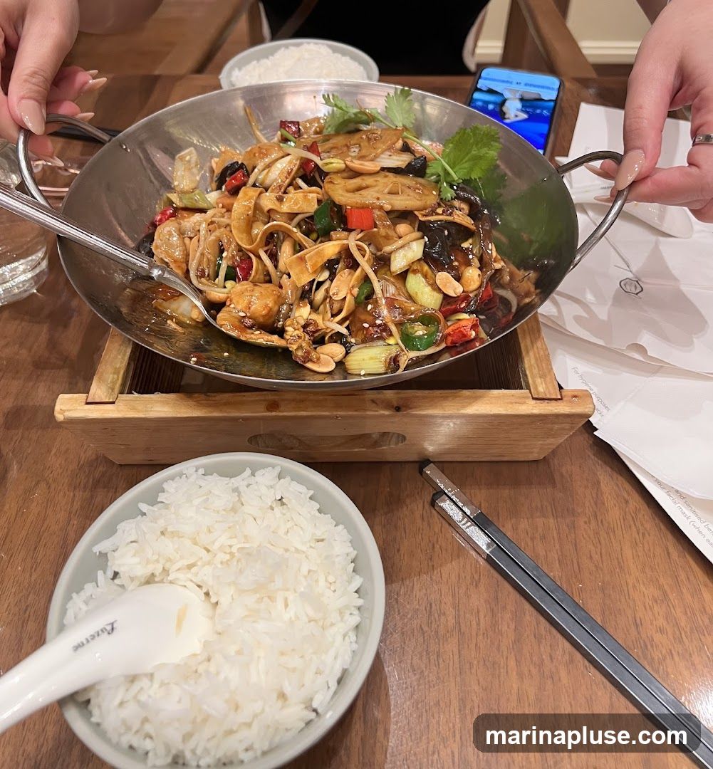 A close-up shot of a table setting with a bowl of white rice and a metal dish filled with a stir-fry dish. The stir-fry appears to contain noodles, lotus root slices, peanuts, chili peppers, and green onions, all coated in a dark, glossy sauce. A spoon rests in the bowl of rice, and chopsticks are placed to the side. The meal is presented on a wooden table, with hands visible at the top and right of the frame, suggesting diners. A phone is also visible in the background.