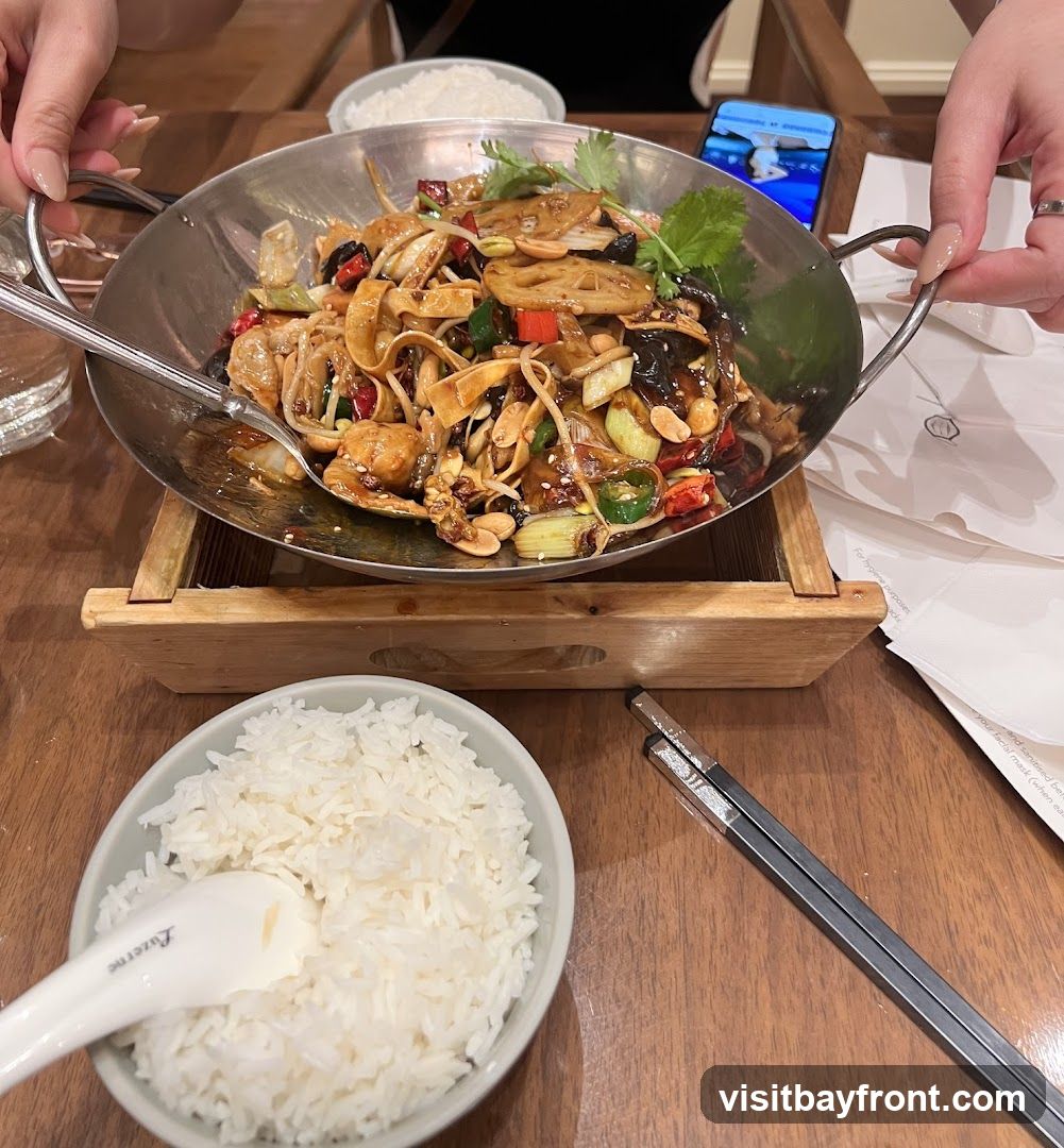 A close-up shot of a table setting with a bowl of white rice and a metal dish filled with a stir-fry dish. The stir-fry appears to contain noodles, lotus root slices, peanuts, chili peppers, and green onions, all coated in a dark, glossy sauce. A spoon rests in the bowl of rice, and chopsticks are placed to the side. The meal is presented on a wooden table, with hands visible at the top and right of the frame, suggesting diners. A phone is also visible in the background.
