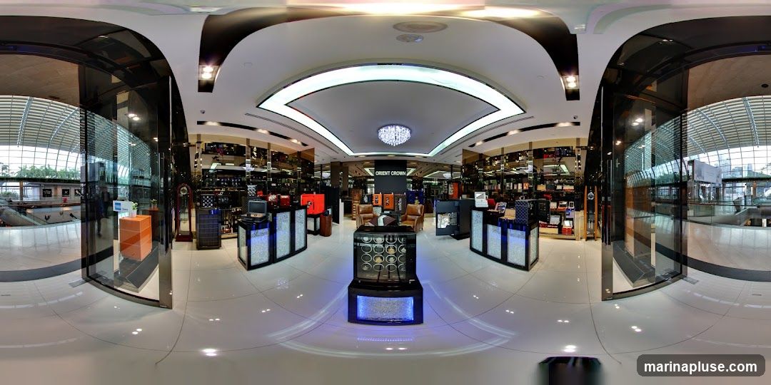 A panoramic view of the interior of a luxury watch store. The store features sleek, modern displays with glass cases showcasing watches. In the center, a prominent sign reads "ORIENT CROWN." There are also comfortable seating areas with leather chairs. The store is well-lit with a chandelier and ambient lighting. The exterior of the mall or shopping center is visible through large glass windows, showing a modern architectural design with a curved roof and natural light.