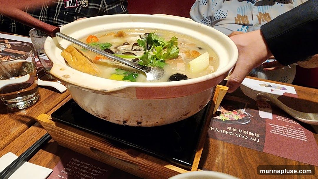 A close-up view of a hot pot dish served in a ceramic pot on a wooden stand. The pot contains a light-colored broth with various ingredients including tofu, mushrooms, and greens. A ladle is placed in the pot. Several people are visible in the background, and a menu or promotional flyer is on the table.