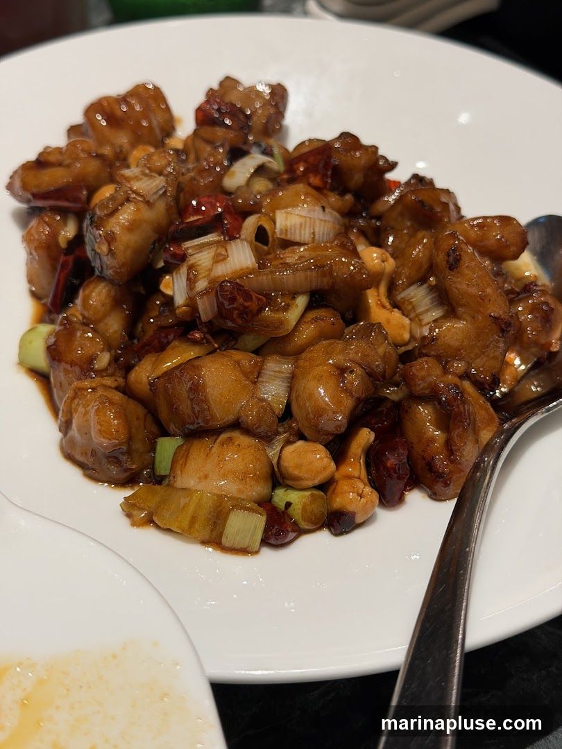 A close-up shot of Kung Pao Chicken served on a white plate. The dish features bite-sized pieces of chicken coated in a glossy, dark sauce, mixed with peanuts, dried chili peppers, and chopped scallions. A fork is visible on the right side of the plate.