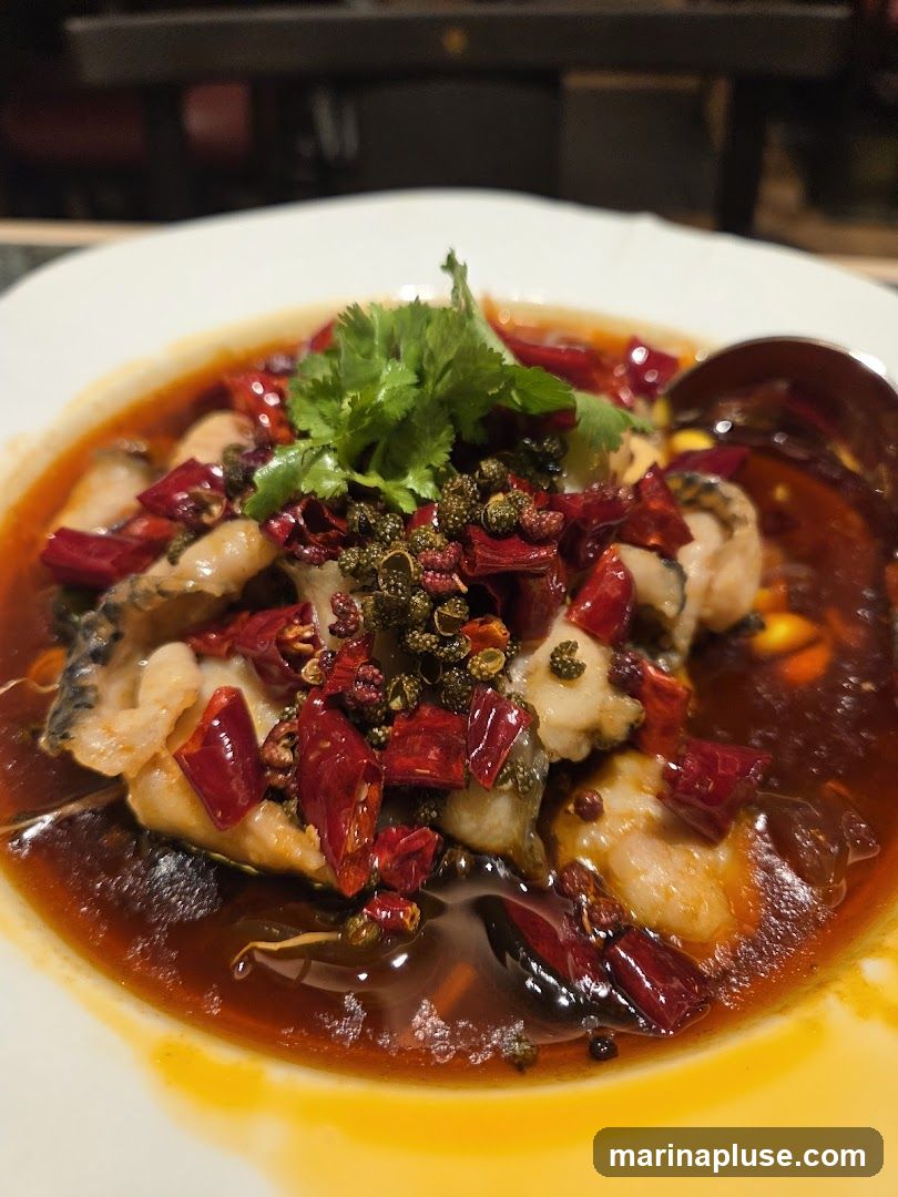 A close-up shot of a white bowl filled with a spicy fish dish. The fish pieces are submerged in a rich, red chili oil broth, generously topped with dried red chili peppers and Sichuan peppercorns. A sprig of fresh cilantro garnishes the top. A spoon is visible on the right side of the bowl. The background is blurred, suggesting an indoor dining setting.