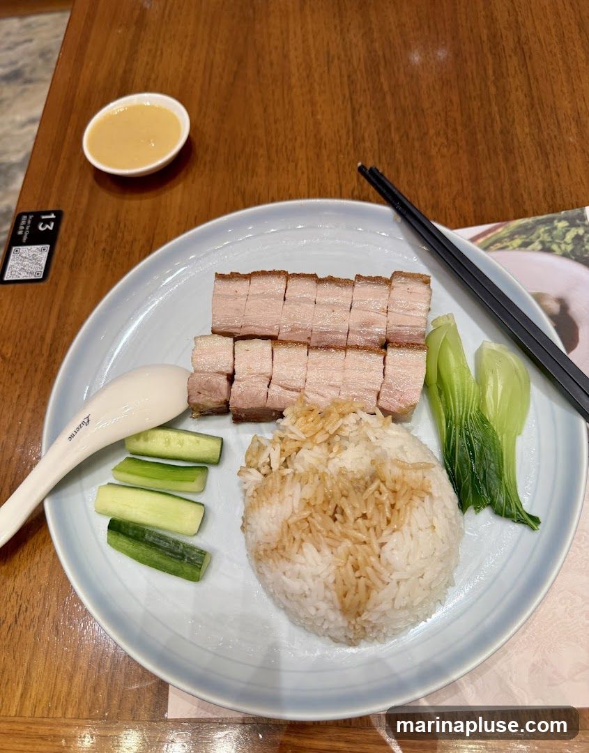 A plate of sliced roast pork belly with crispy skin, served with a mound of white rice drizzled with sauce, cucumber slices, and blanched greens. A small dish of dipping sauce is also on the table, along with chopsticks and a spoon. A table number tag is visible in the upper left corner.