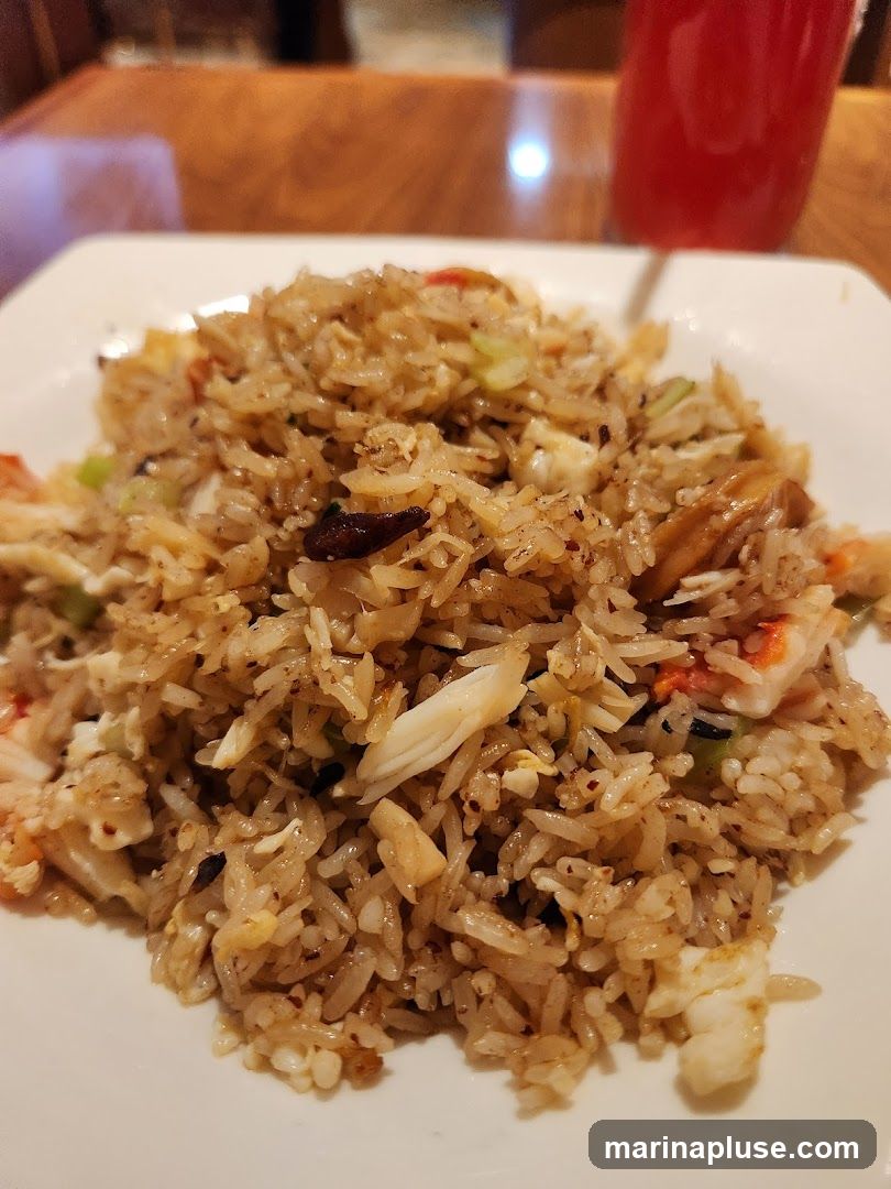 A close-up shot of crab fried rice on a white plate. The rice is mixed with pieces of crab meat, scrambled egg, and green onions. In the background, a red drink in a glass and a wooden table are visible.