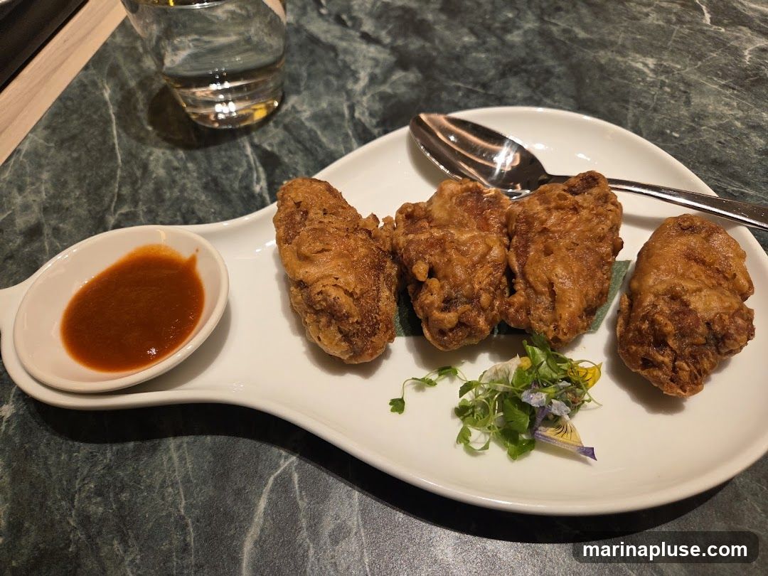 A plate of golden-brown fried chicken pieces served with a small dish of red dipping sauce and a garnish of microgreens and edible flowers. A spoon rests on the plate, and a glass of water is visible in the background.