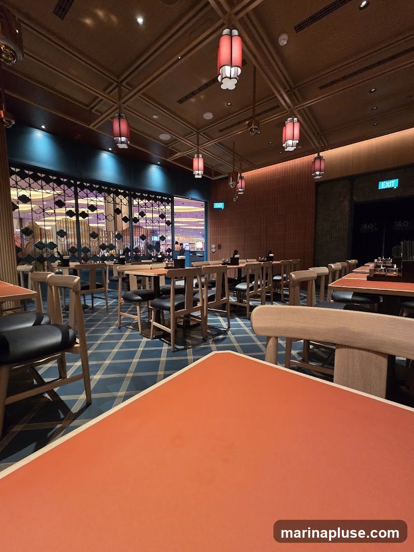 The image shows the interior of a restaurant with a modern design. The ceiling features a wooden lattice pattern with red lanterns hanging down. The walls are a mix of exposed brick and dark paneling. The floor is covered in a blue and gold checkered pattern. Tables are set with orange placemats and wooden chairs with black seats. In the background, through a decorative screen, a brightly lit area, possibly a casino or entertainment venue, is visible. Exit signs are present on the walls.