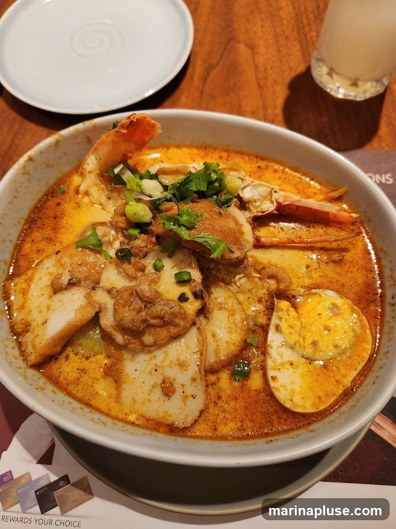 A close-up shot of a bowl of seafood laksa, a spicy noodle soup. The dish features a rich, orange broth, topped with a large prawn, crab pieces, sliced fish cakes, and a hard-boiled egg cut in half. Garnished with chopped green onions and cilantro. A small white plate and a glass of a light-colored beverage are visible in the background. A placemat with credit card logos and the text "REWARDS YOUR CHOICE" is partially visible at the bottom.