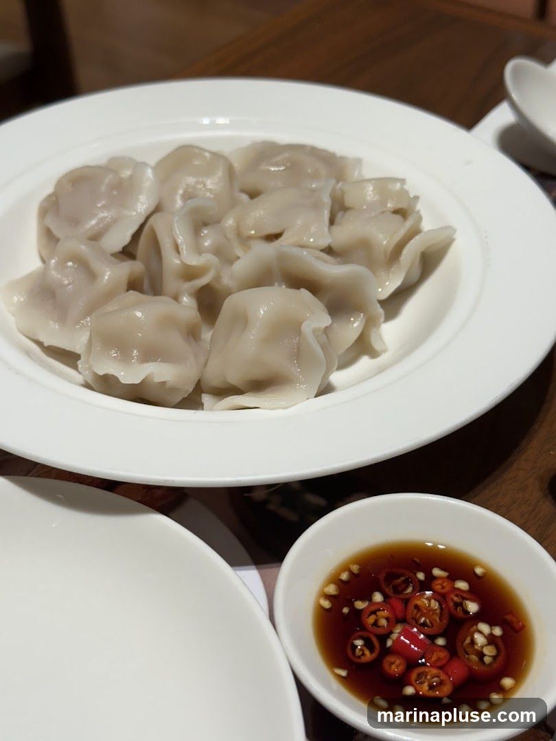 A white bowl filled with steamed dumplings sits on a wooden table. To the right, a small white dish contains a dark dipping sauce with sliced red chilies and seeds. Another white plate is partially visible in the foreground.