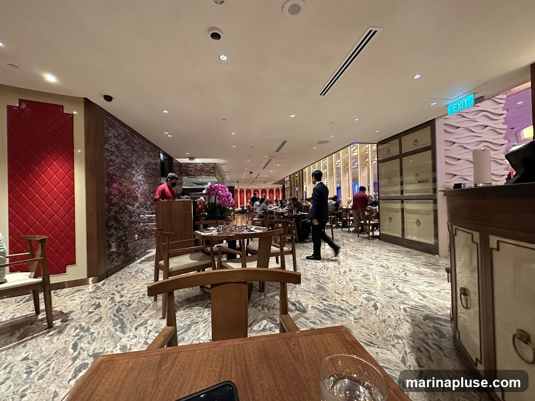 An interior view of a restaurant with tables and chairs set up for dining. Several patrons are seated at tables, and staff members are visible. The decor includes patterned walls, wooden furniture, and decorative elements. An 'EXIT' sign is illuminated in green.
