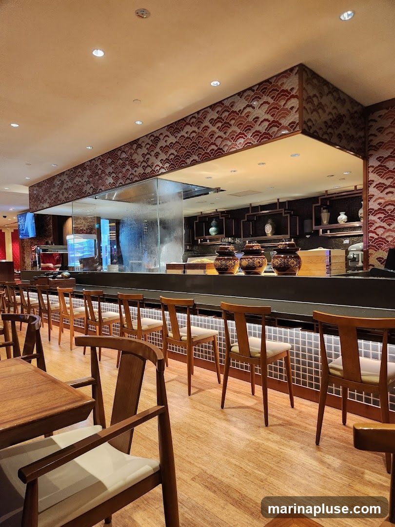 The image shows the interior of a restaurant with a long bar area. Several wooden chairs with light-colored cushions are neatly arranged along the bar. The bar itself has a dark countertop and a tiled backsplash illuminated by soft lighting. Above the bar, decorative panels with a wave-like pattern are visible. In the background, shelves display various ceramic vases and jars. The flooring is made of polished wood planks. Tables and chairs are also visible in the foreground, suggesting a dining area.