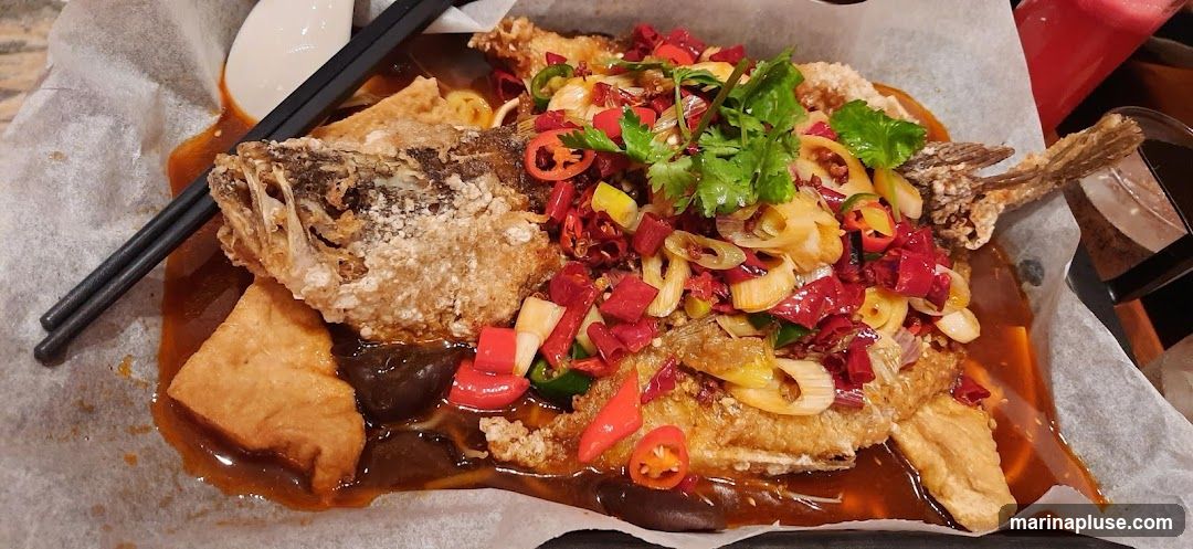 A whole fried fish is served in a spicy sauce with chili peppers, scallions, cilantro, and pieces of fried tofu. Chopsticks are resting on the dish.