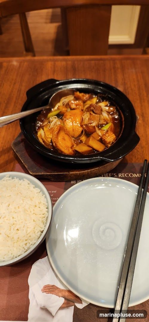 A close-up shot of a braised dish served in a black pot, accompanied by a bowl of white rice and a plate with chopsticks. The dish appears to contain meat, possibly abalone, and vegetables in a rich brown sauce. The setting is a restaurant with wooden furniture. A placemat with text and an image of a chef is visible beneath the plate and rice.