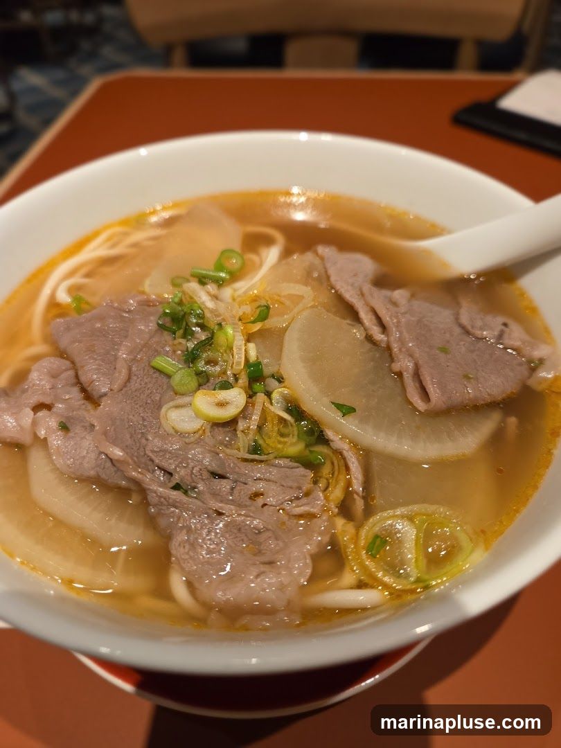 A close-up shot of a bowl of beef noodle soup. The soup is a rich orange broth with thin noodles, slices of beef, and chunks of white radish. It is garnished with chopped green onions and what appears to be fried shallots. A white spoon rests in the bowl. The background is blurred, showing a reddish-brown table and hints of a dark blue patterned surface.