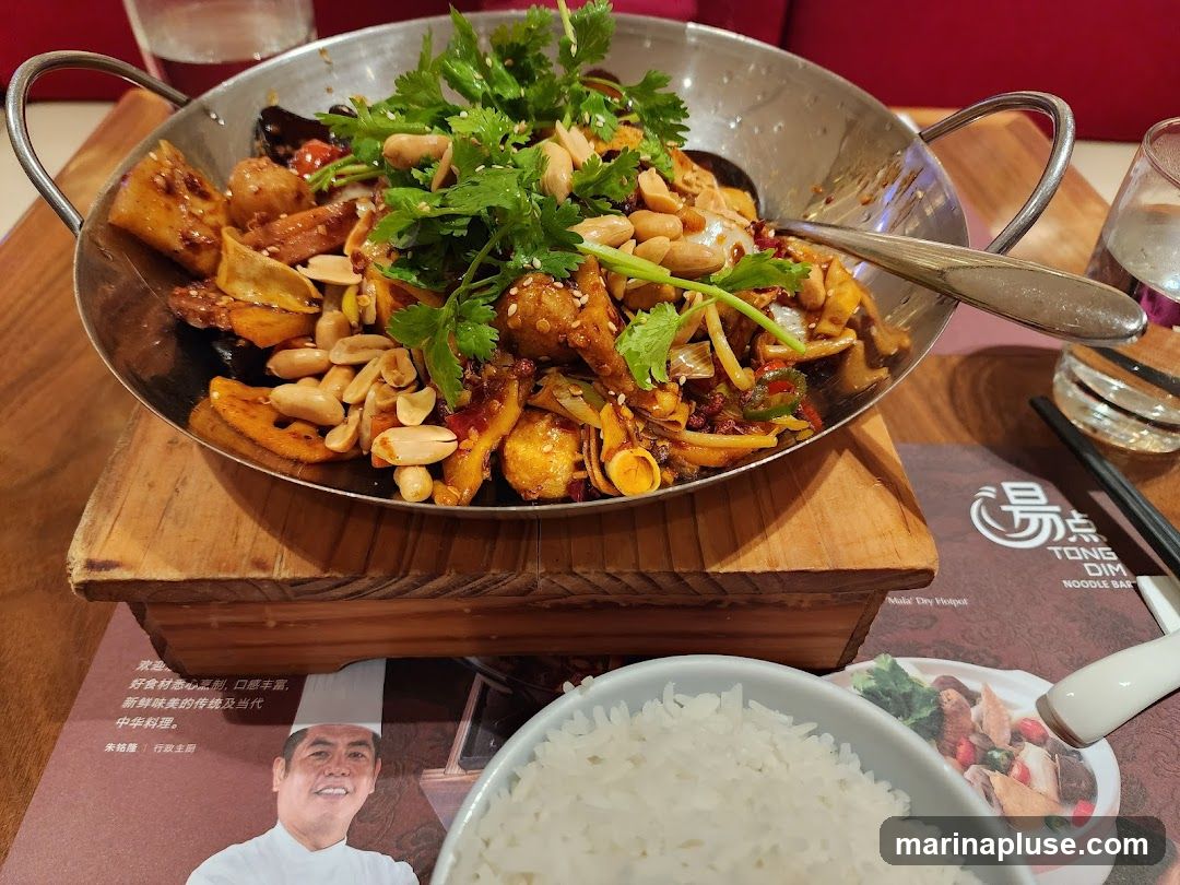 A close-up shot of a spicy stir-fry dish served in a metal wok, topped with peanuts and fresh cilantro. The dish contains various ingredients including what appears to be tofu, vegetables, and chili peppers. A bowl of white rice is visible in the foreground, and a menu with Chinese text and the logo "TONG DIM NOODLE BAR" is partially visible beneath the wok. A glass of water and chopsticks are also present.