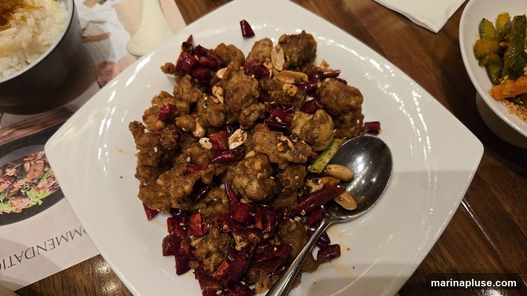 A plate of crispy fried chicken pieces seasoned with dried chili peppers and peanuts is served with a side of rice and pickled vegetables. A spoon rests on the plate.