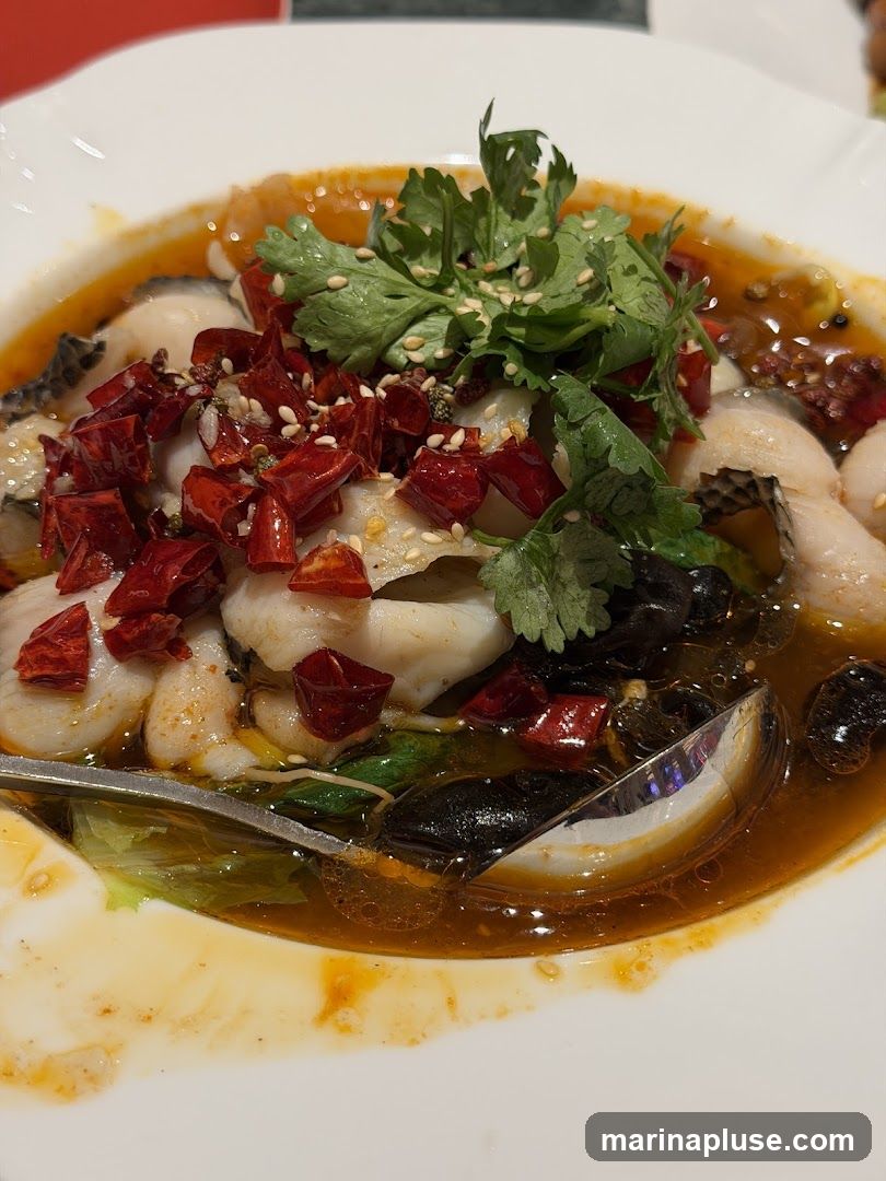 A close-up shot of a white bowl filled with a spicy fish dish. The dish features white fish fillets, dried red chilies, cilantro, sesame seeds, and what appear to be black mushrooms and some leafy greens. The fish is submerged in a reddish-orange broth. A spoon is visible in the lower right of the bowl.