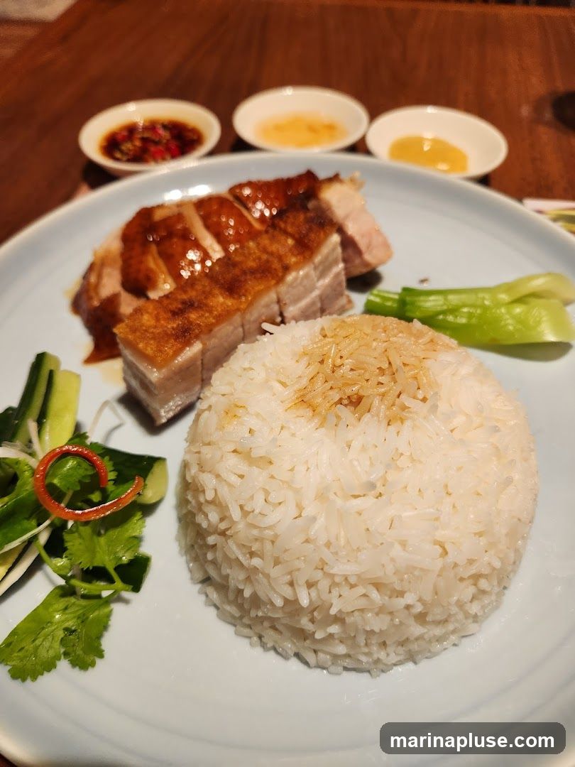 A plate of white rice is served with slices of roast pork and crispy pork belly. The dish is garnished with cucumber slices, cilantro, and a swirl of red pepper. Three small dipping sauces are arranged in the background.