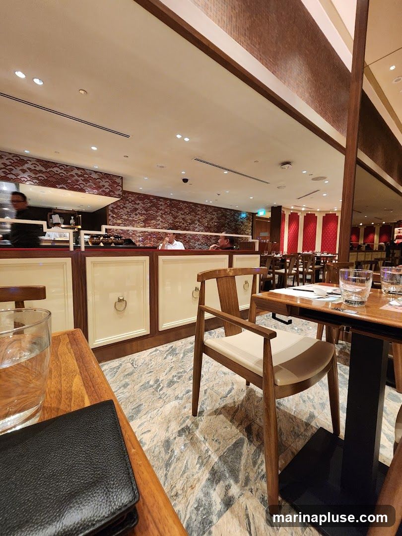 A view of a restaurant interior with tables and chairs. In the background, chefs can be seen working behind a counter. The decor features red and white patterned wallpaper and red upholstered panels on the walls.