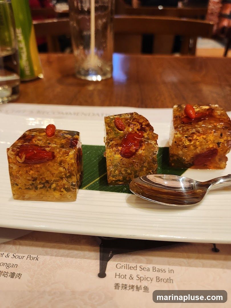 Three cubes of jelly-like food with goji berries and nuts are served on a white plate with a spoon. A menu is visible below the plate.