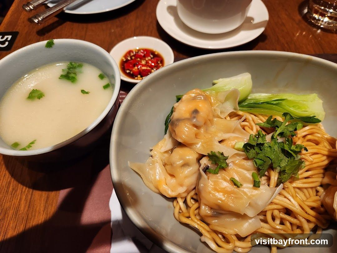 A close-up shot of a bowl of wonton noodles, garnished with fresh cilantro and bok choy. A side of soup and a small dish of chili sauce are also visible.