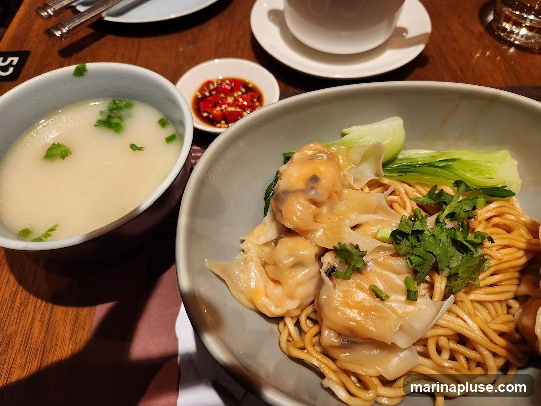 A close-up shot of a bowl of wonton noodles, garnished with fresh cilantro and bok choy. A side of soup and a small dish of chili sauce are also visible.