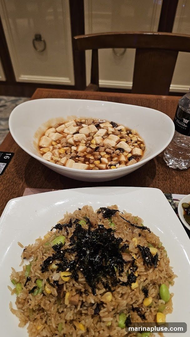 A close-up shot of two dishes on a wooden table. In the foreground, a white plate holds a serving of fried rice topped with shredded seaweed. The fried rice contains edamame and corn. Behind it, a white bowl contains a dish of tofu and what appears to be minced meat in a savory sauce. A bottle of water with a label that reads "Marina Bay Sands Singapore" is visible to the right. The background shows a portion of a wooden chair and decorative wall panels.