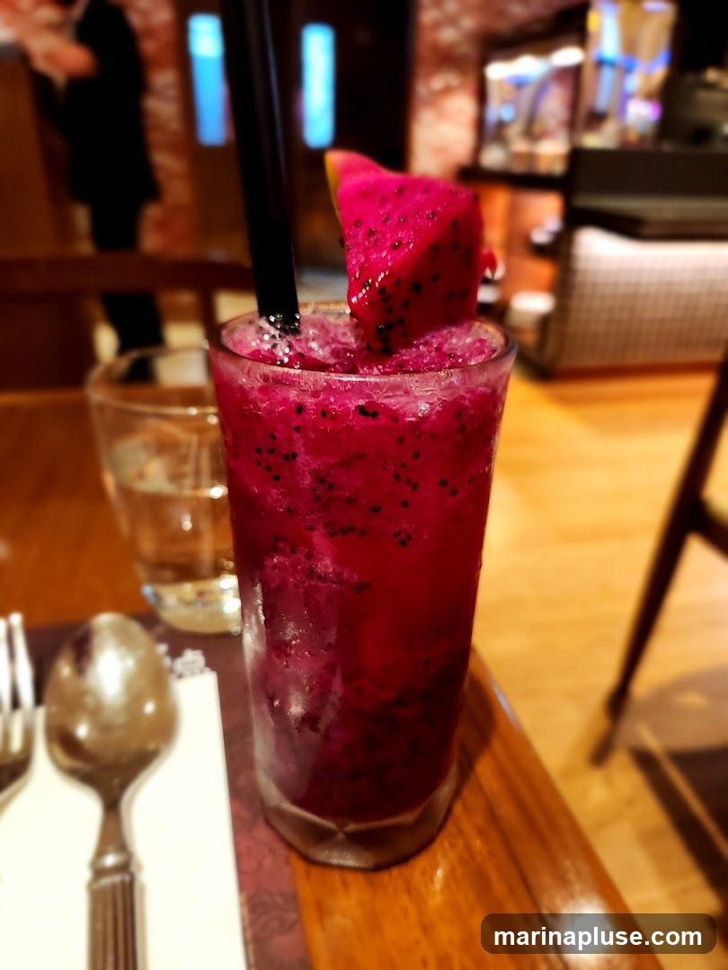 A close-up shot of a vibrant pink dragon fruit smoothie in a tall glass, garnished with a slice of dragon fruit and a black straw. The drink is filled with crushed ice and has visible black seeds from the fruit. In the background, a blurred image shows a person in silhouette and a dimly lit interior with what appears to be a bar or counter area. A glass of water and cutlery are visible on the left side of the frame, suggesting a dining setting.