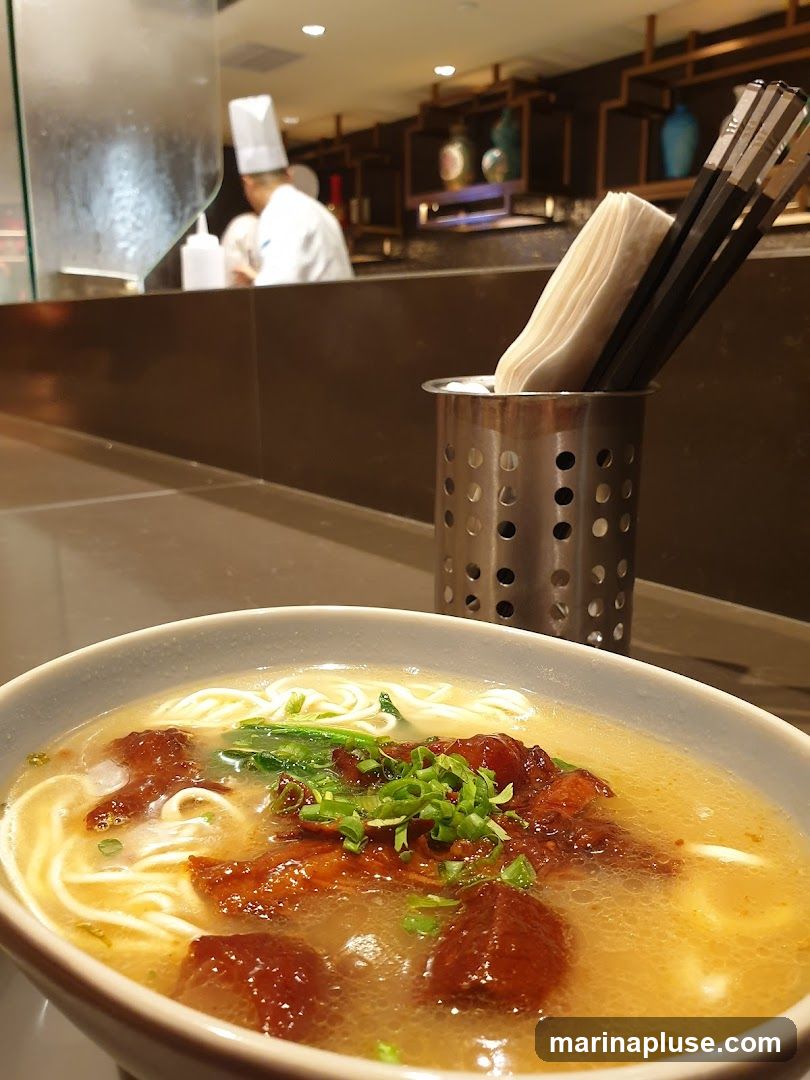 A close-up view of a bowl of noodle soup with chunks of meat and green onions. In the background, a chef in a white uniform is visible behind a counter, and shelves with decorative items are on the wall. Chopsticks and napkins are in a metal holder on the counter.