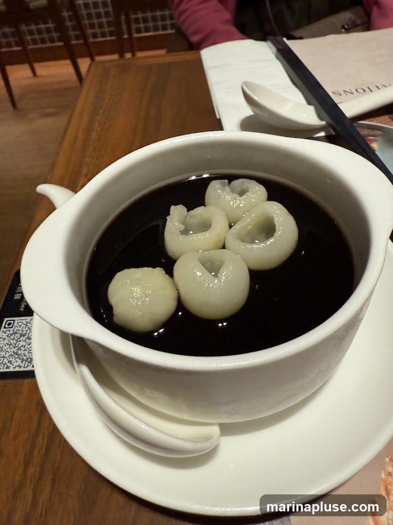 A white bowl filled with a dark liquid dessert, topped with several lychees. The bowl sits on a saucer with a spoon. In the background, a wooden table, chairs, and a menu are visible.