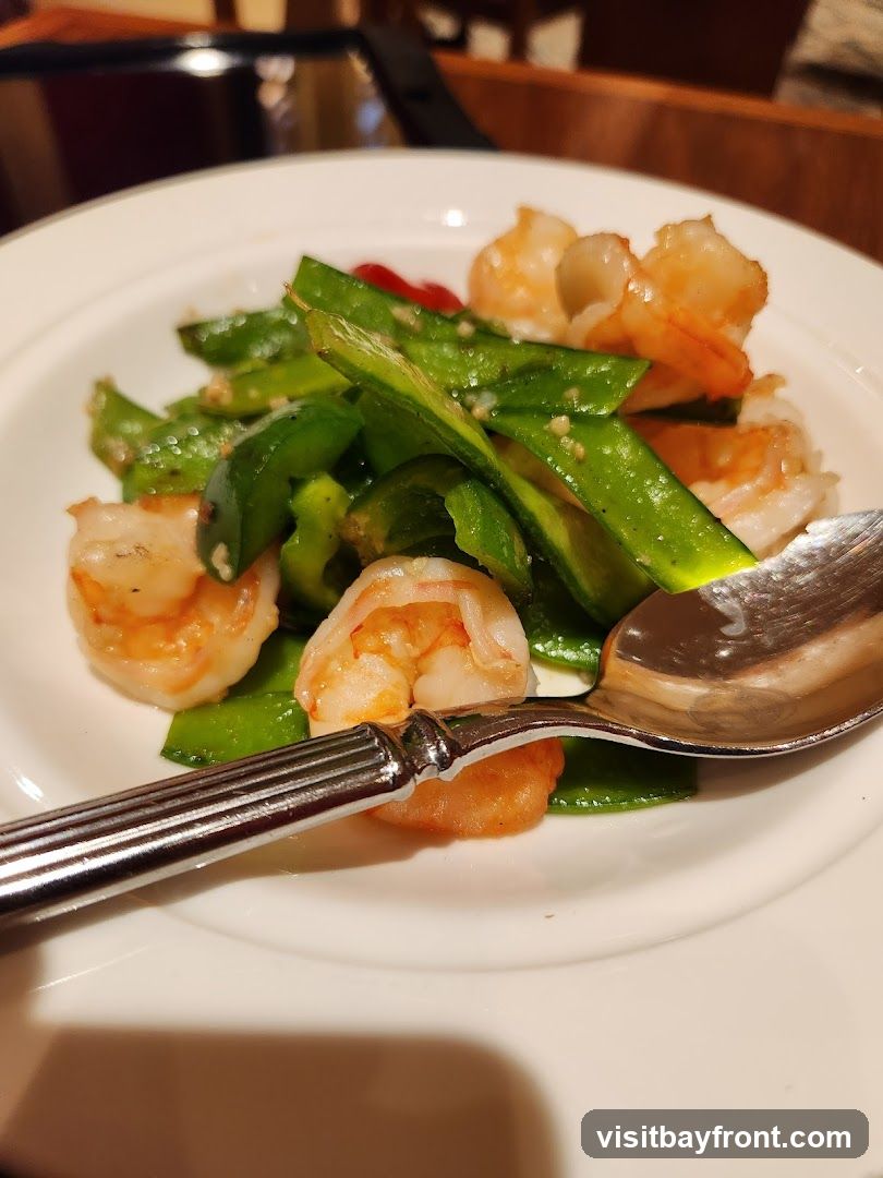 A close-up shot of a white plate filled with a dish of shrimp and green bell peppers. A spoon rests on the side of the plate. The dish appears to be seasoned with small flecks of spices.