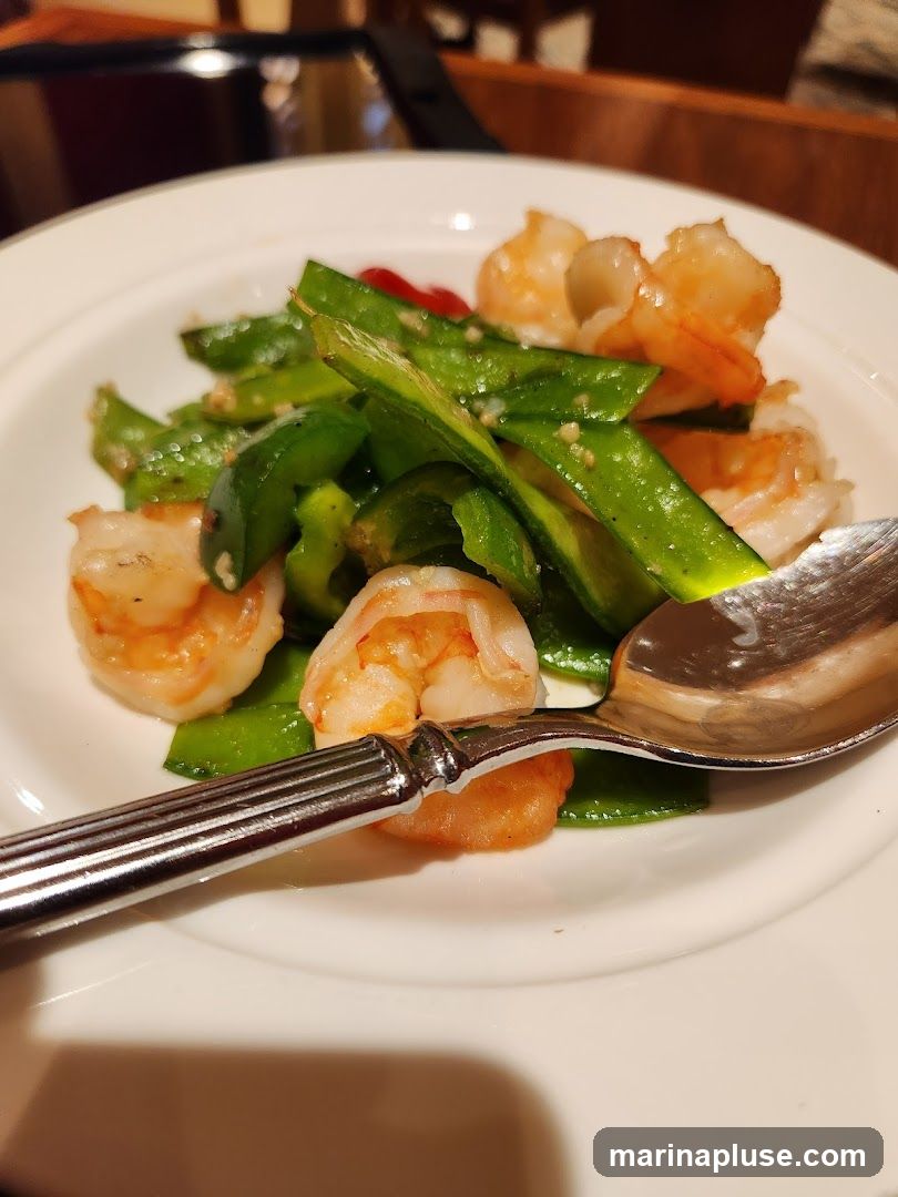 A close-up shot of a white plate filled with a dish of shrimp and green bell peppers. A spoon rests on the side of the plate. The dish appears to be seasoned with small flecks of spices.