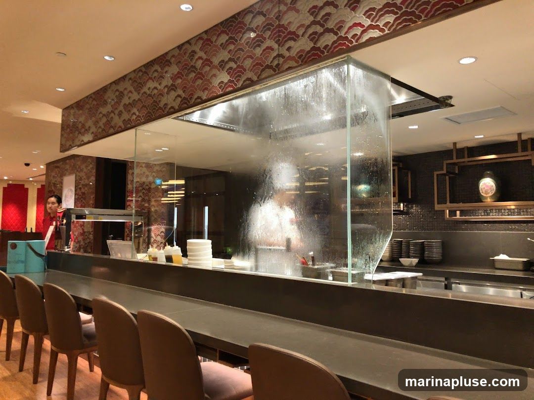 A view of a restaurant kitchen counter with a glass partition. A chef is visible working behind the glass. There are chairs lined up in front of the counter. The decor includes patterned wallpaper and decorative shelving.
