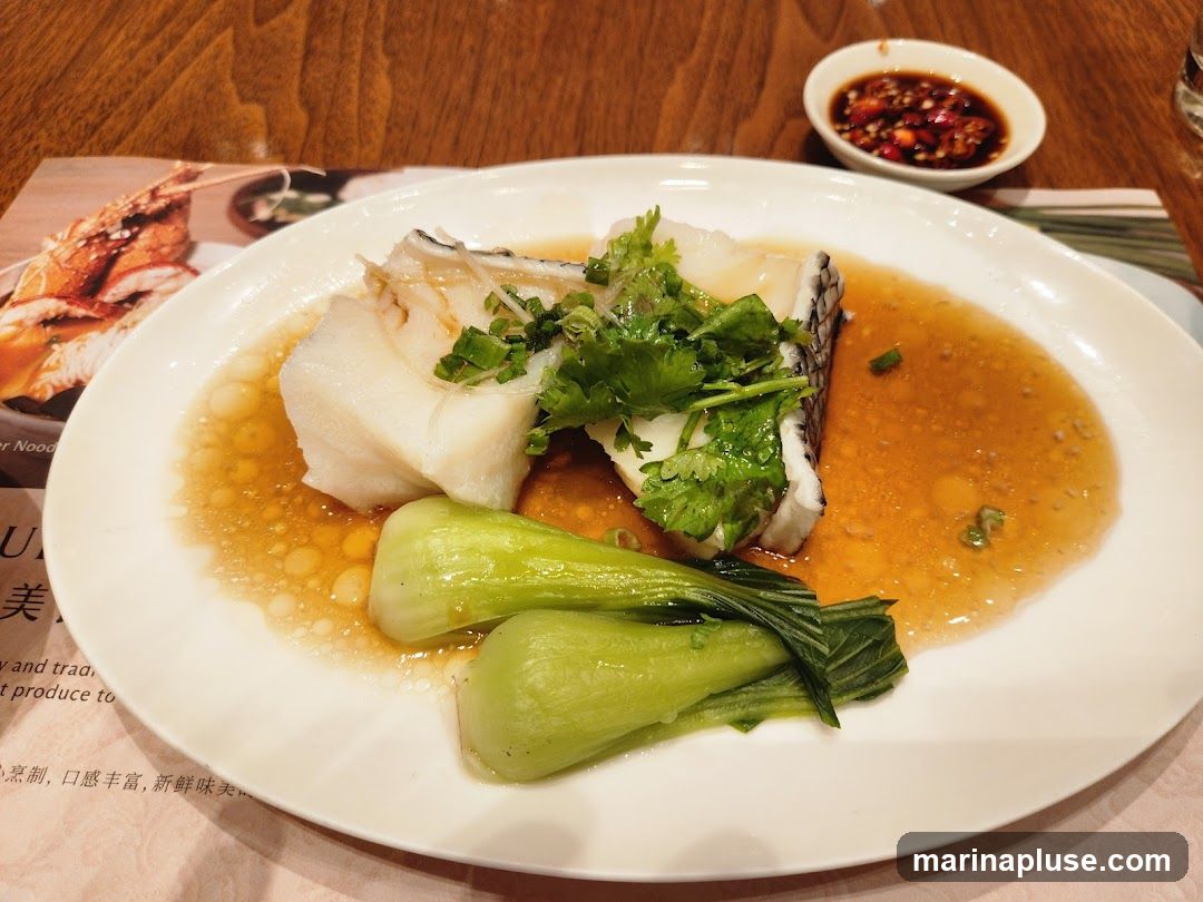 A white plate holds a serving of steamed fish, garnished with cilantro and green onions, and served with bok choy. A small dish of dipping sauce is to the side. A menu with images of food is visible in the background.