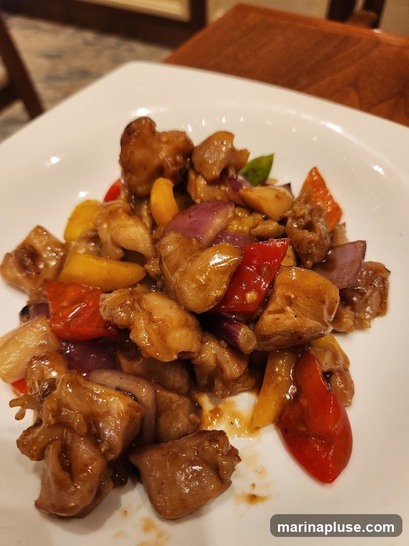 A close-up shot of a white plate filled with a stir-fry dish. The dish contains bite-sized pieces of chicken, red and yellow bell peppers, and red onions, all coated in a glossy brown sauce. The food is presented on a white, square-edged plate, with a blurred background suggesting a dining setting with wooden furniture.