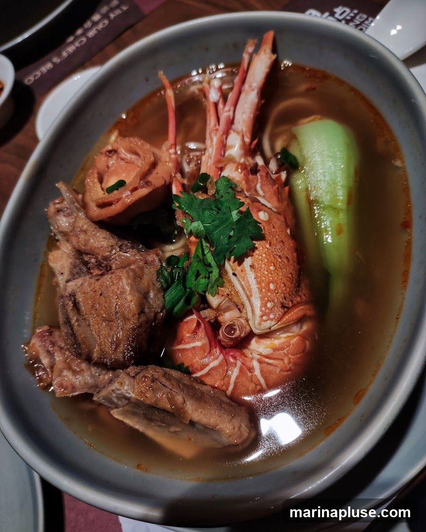 A close-up shot of a bowl of noodle soup. The soup contains a lobster, pieces of meat, noodles, bok choy, and cilantro. The broth is a reddish-brown color.