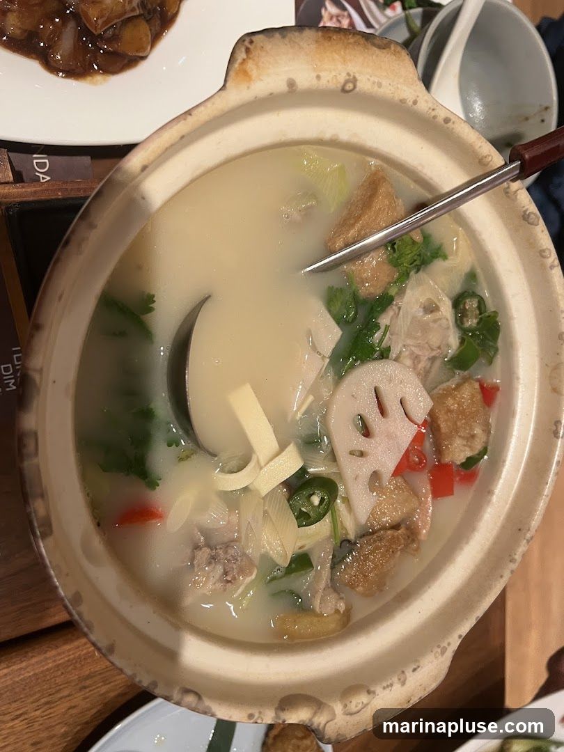 A close-up overhead view of a steaming hot pot soup in a ceramic pot. The soup is a milky white broth filled with various ingredients including pieces of chicken, tofu, lotus root slices, green onions, and cilantro. A ladle is partially submerged in the soup, and a metal serving spoon rests on the edge of the pot. In the background, another dish with dark sauce and food is visible, along with a white bowl and spoon.