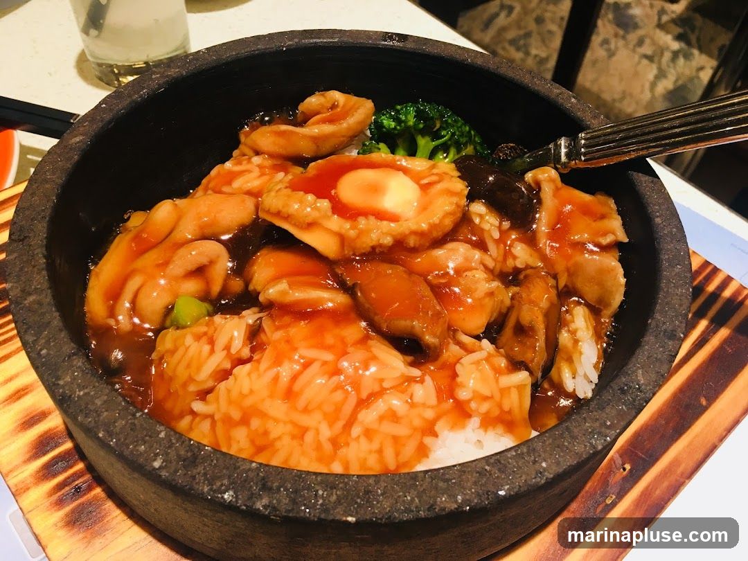 A close-up shot of a stone bowl filled with rice and topped with a savory sauce, abalone, mushrooms, and broccoli. A fork rests on the edge of the bowl.