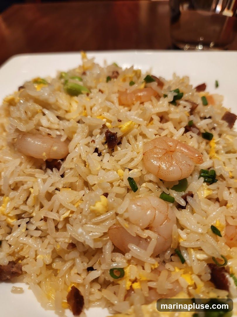 A close-up shot of a plate of shrimp fried rice. The rice is mixed with scrambled eggs, small pieces of meat, green onions, and several pink shrimp. The dish is served on a white plate.