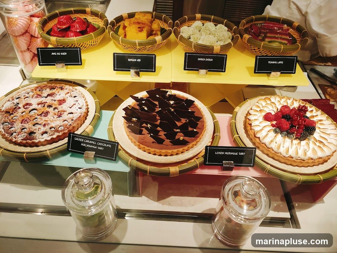 A display of various desserts including tarts, cakes, and traditional sweets. Visible items include Ang Ku Kueh, Bingka Ubi, Ondeh Ondeh, Penang Lapis, Salted Caramel Chocolate Macadamia Tart, and Lemon Meringue Tart. There are also meringues and jars of candies.