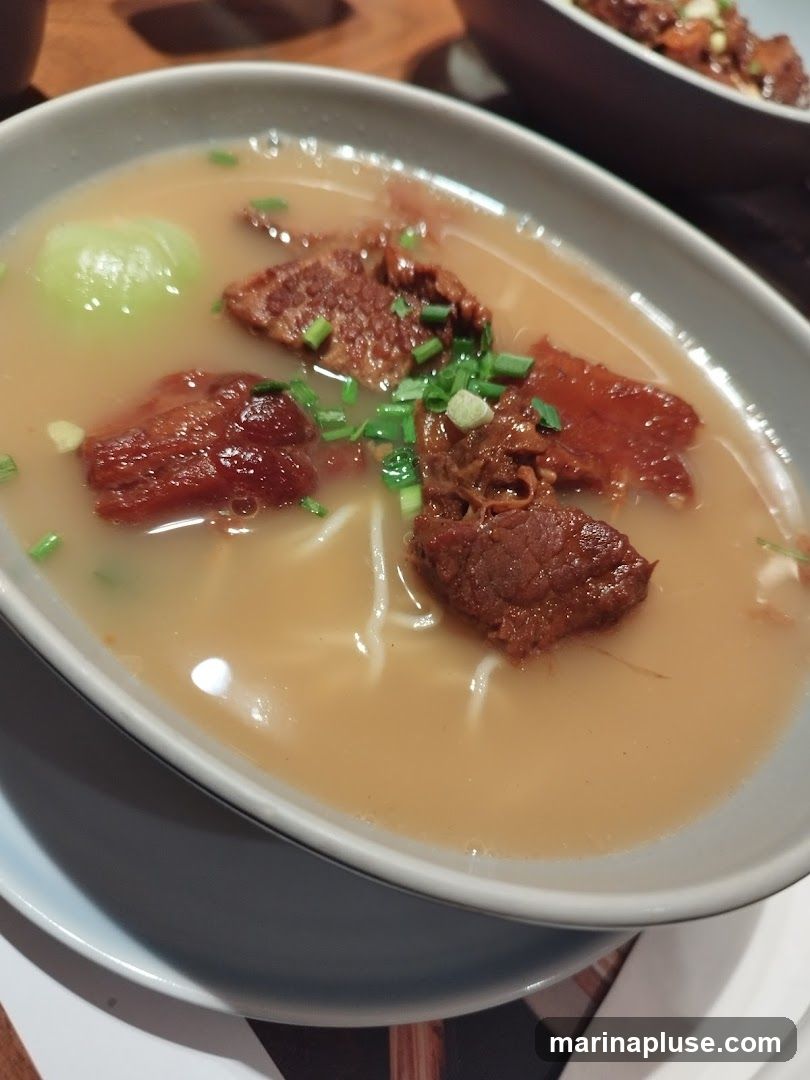 A close-up shot of a bowl of beef noodle soup. The soup is a light brown color and contains several pieces of tender-looking beef, noodles, and a piece of green vegetable, possibly bok choy. Chopped green onions are sprinkled on top. Another bowl of food is visible in the blurred background.