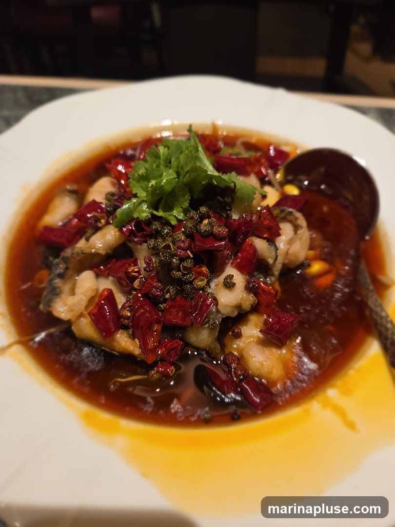 A close-up shot of a white plate filled with a dish of fish in a spicy, oily sauce. The dish is garnished with fresh cilantro, dried red chili peppers, and Sichuan peppercorns. A dark spoon rests on the right side of the plate. The background is blurred, suggesting an indoor dining setting.