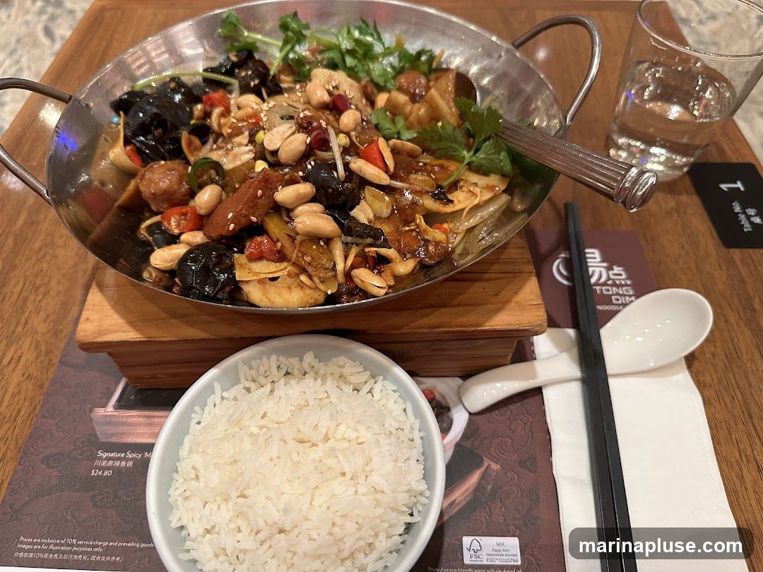 A close-up shot of a dish of spicy stir-fried food served in a metal bowl, accompanied by a bowl of white rice. The stir-fry contains various ingredients such as peanuts, mushrooms, and what appear to be meat pieces, all coated in a glistening sauce. A glass of water, chopsticks, and a spoon are also visible on the table. A menu or placemat with the restaurant's logo and some text is partially visible.