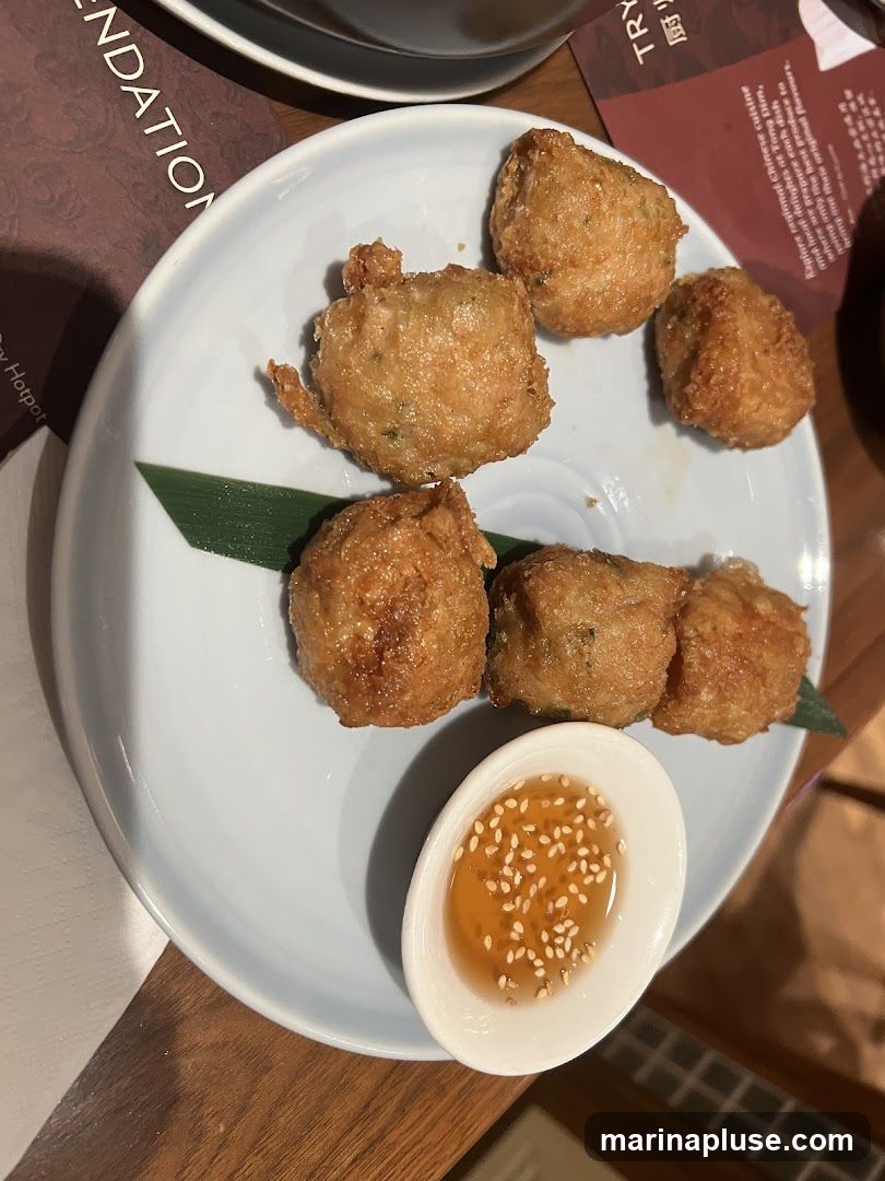 A plate of golden-brown fried appetizers, possibly shrimp cakes or meatballs, is served with a small dish of dipping sauce sprinkled with sesame seeds. A green leaf garnishes the plate. In the background, a menu or placemat with text is visible.