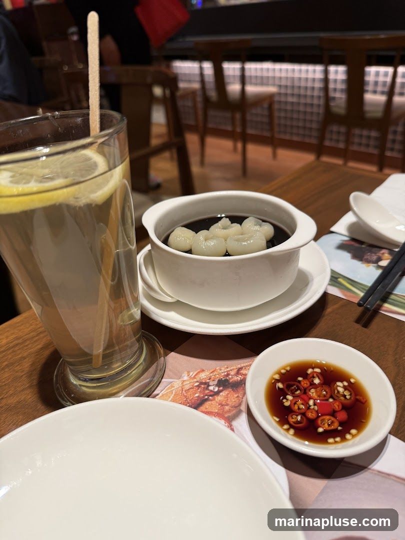 A close-up view of a table setting with a glass of iced lemon tea, a bowl of black jelly dessert with lychee, and a small dish of chili sauce. The background shows a dimly lit restaurant interior with wooden chairs and a counter.