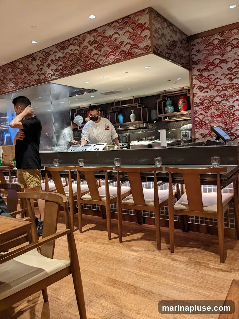A view of a restaurant's open kitchen and dining area. Chefs are visible working behind a counter, with food preparation stations and equipment. The dining area features wooden tables and chairs, with decorative wallpaper on the walls.