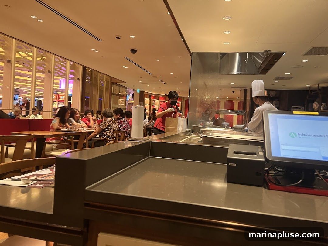 A view inside a restaurant, showing diners seated at tables in the background and chefs working behind a counter in the foreground. One chef is wearing a tall white hat and appears to be cooking, while another, also in a chef's hat, is handing a bag to someone. A computer monitor and a receipt printer are visible on the counter. The restaurant has a modern interior with warm lighting.