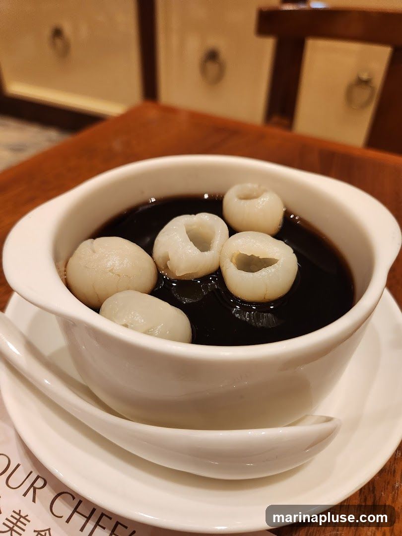 A white bowl filled with dark grass jelly and topped with several peeled lychees. The bowl sits on a white saucer with a white spoon. A portion of a menu with Chinese characters and the words "OUR CHEF" is visible in the lower left corner. The background shows a wooden table and the backs of chairs.