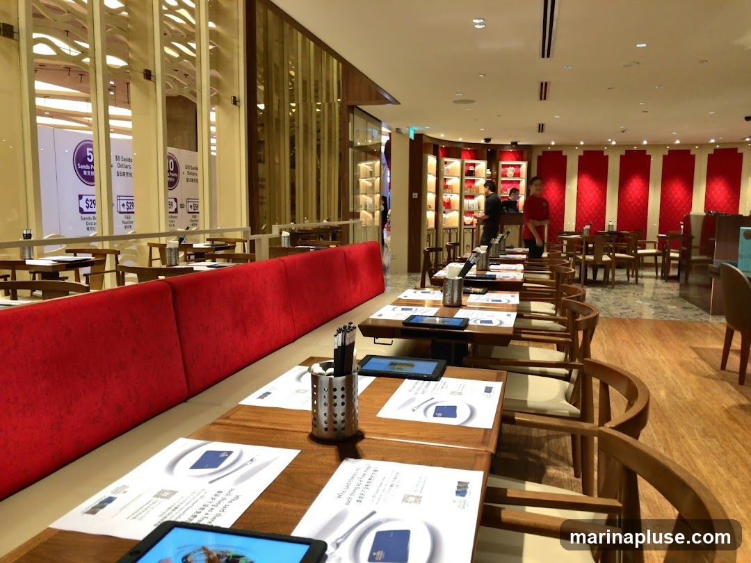 The image shows the interior of a restaurant with wooden tables and chairs. Red banquettes line one side of the dining area. In the background, staff members are visible near a counter, and shelves display items. Promotional signs are visible through a glass partition on the left.