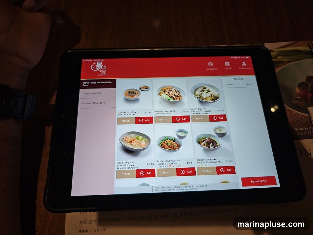 A tablet displays a digital menu for a restaurant called TONG DIM NOODLE BAR. The menu features various noodle dishes and dim sum items with prices and images. Dishes include Sichuan Pork Chop Dan Dan Noodles, Assorted Mushrooms with Dry Noodles, Fish Broth Noodle, Hong Kong Style Prawn Dumping Noodles, Dry Noodles with Mini Abalone & Pork with Mushroom, and Braised Beef Brisket Noodles. The tablet shows a shopping cart with selected items and an option to submit the order. A printed menu or flyer with Chinese text and the word "Wok-Fried" is partially visible to the right.